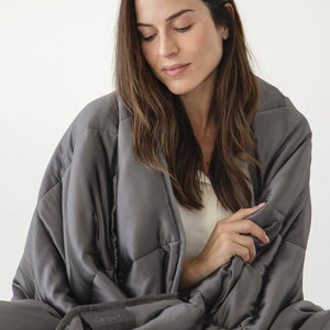 A woman with long brown hair sits wrapped in the drowsysleepco Moonlight Shadow Silk Weighted Blanket, looking down gently against a plain light background.