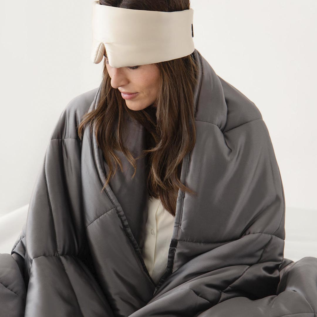 A woman wearing a cream-colored sleep mask and wrapped in a gray weighted blanket sits calmly, looking down with a peaceful expression.