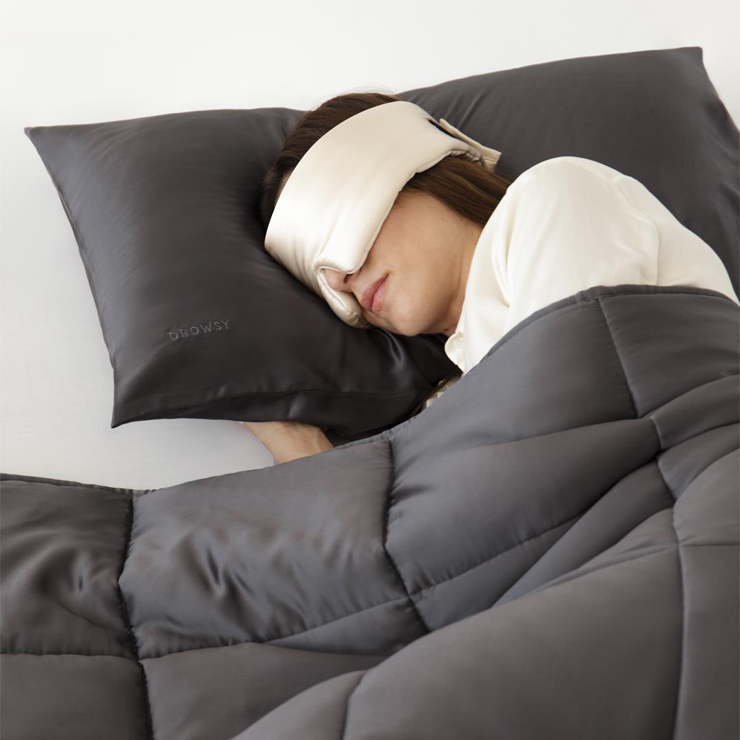 A person sleeps under the drowsysleepco Moonlight Shadow Silk Weighted Blanket in gray, resting their head on a gray pillow, wearing a white eye mask and a white long-sleeve shirt.