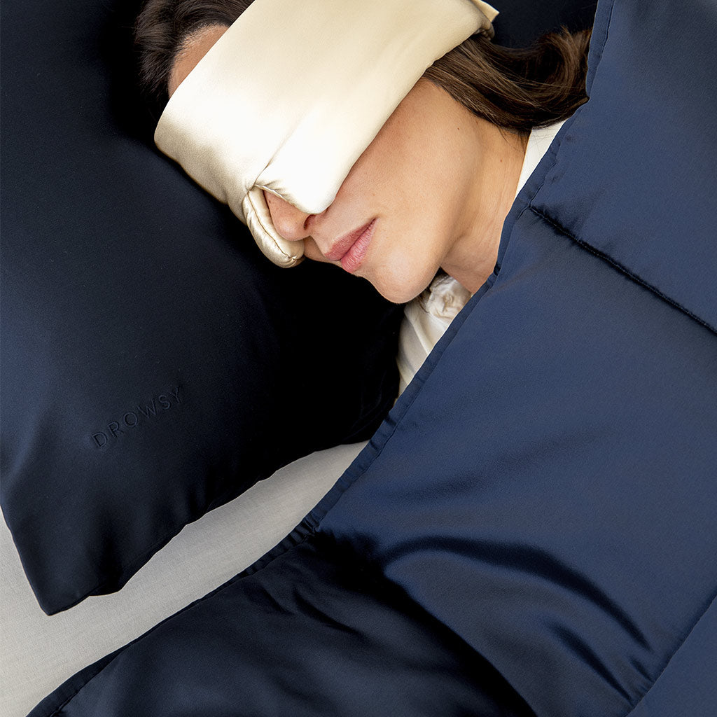 A person lies in bed with a cream-colored sleep mask covering their eyes, resting on a dark blue pillow and covered with a matching dark blue blanket.