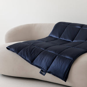 The Midnight Blue Silk Weighted Blanket by drowsysleepco is draped over a modern light beige curved sofa against a white wall, its small black tag visible and inviting deep relaxation.