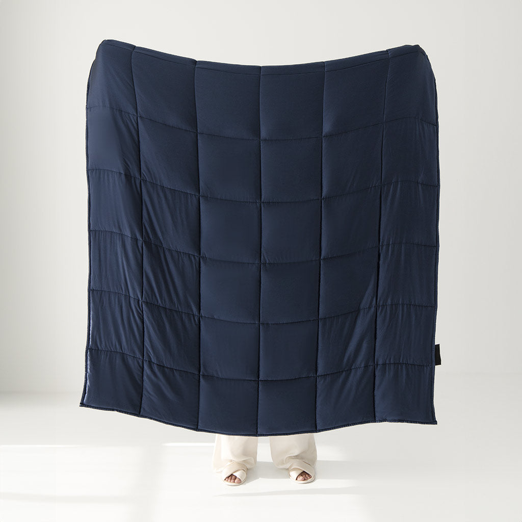 A person stands behind and holds up the drowsysleepco Midnight Blue Silk Weighted Blanket, with only their feet showing. Set against a plain white indoor background, the scene evokes a sense of deep relaxation.