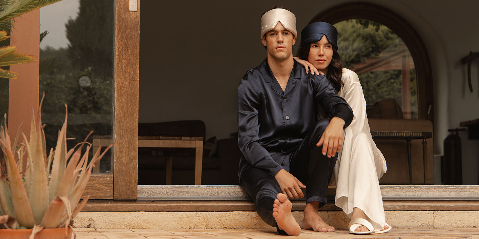 A man and woman in silk pajamas and matching sleep caps sit barefoot on a doorstep. The man wears navy blue and the woman white. They sit close together, looking ahead, with a cozy indoor scene behind them.