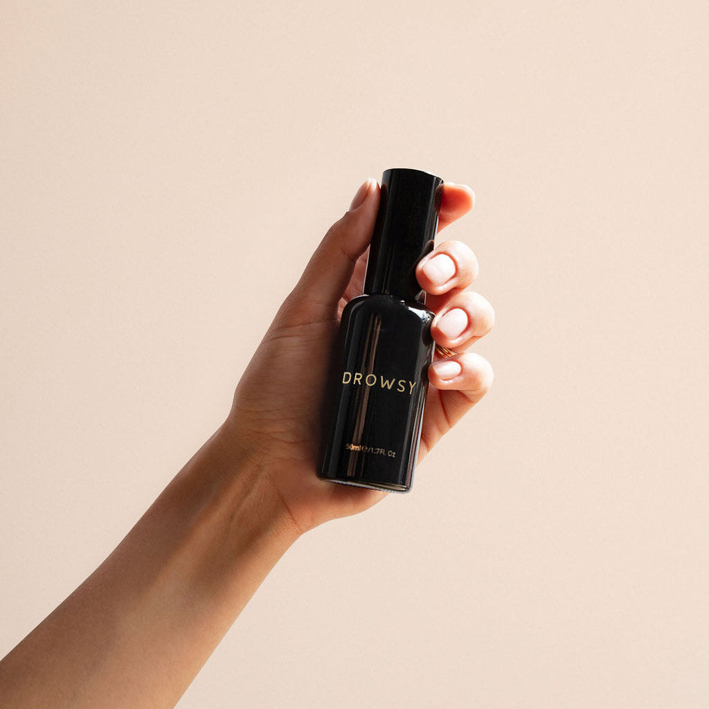 A hand holding a black bottle labeled Sleep S.O.S Pillow Spray by drowsysleepco, a natural sleep aid, against a light beige background.