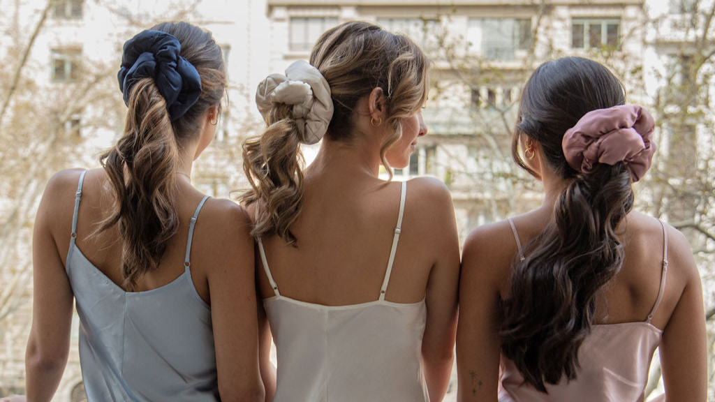Three women with wavy hair wearing satin camisoles and large scrunchies—navy, beige, and pink—stand side by side facing away, looking out a window at city buildings and trees.