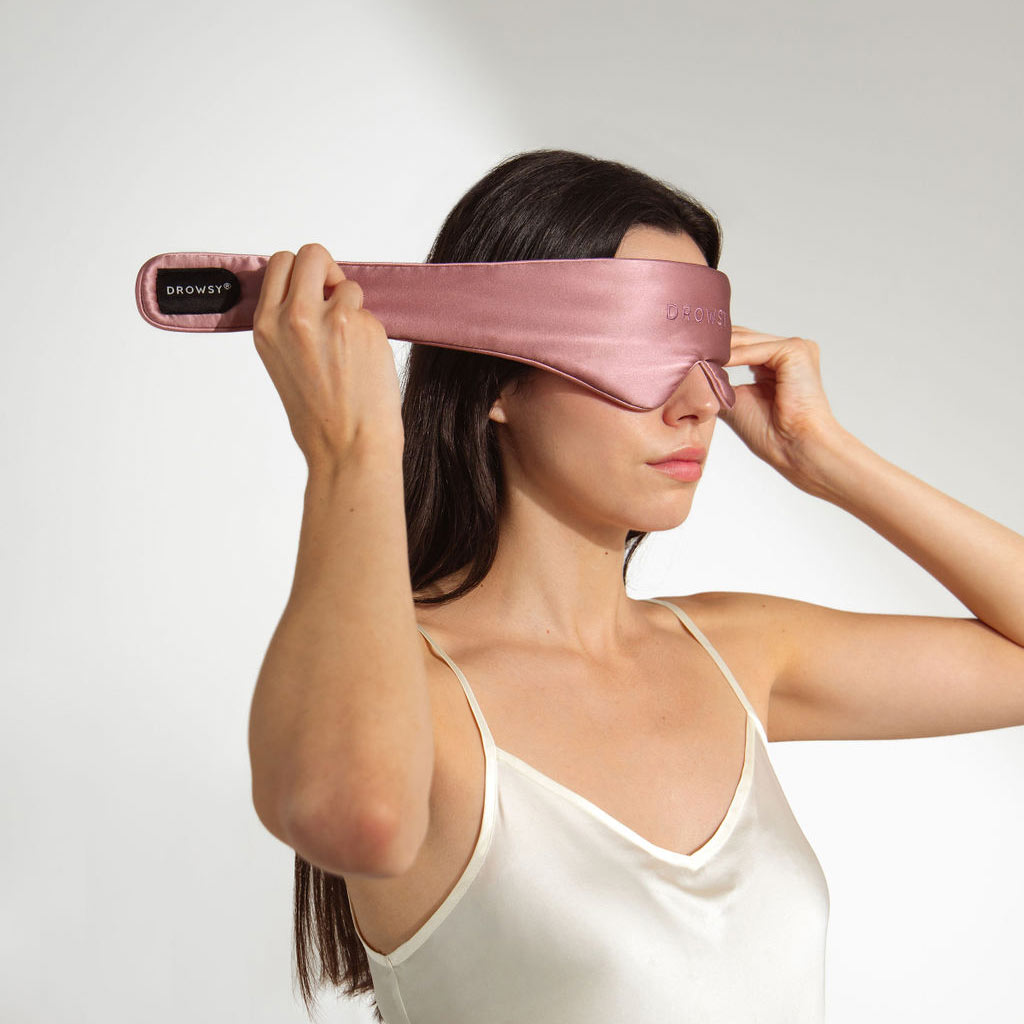 A woman in a white satin camisole puts on the drowsysleepco Damask Rose Featherweight Silk Sleep Mask, holding the strap near her ear. This luxurious mask is perfect for restful sleep against a plain white background.