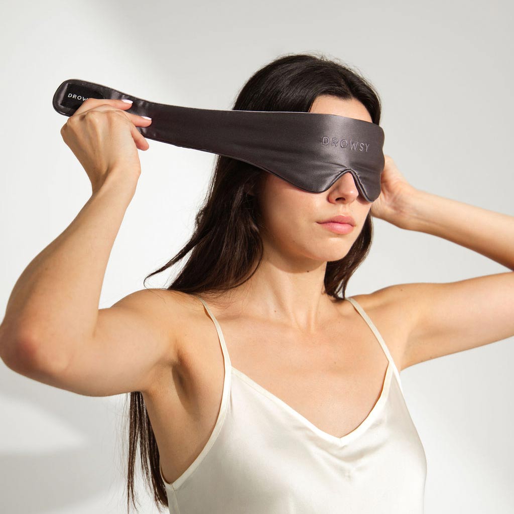 A woman in a white satin camisole adjusts a black sleep mask labeled DROWSY over her eyes, standing against a plain white background.