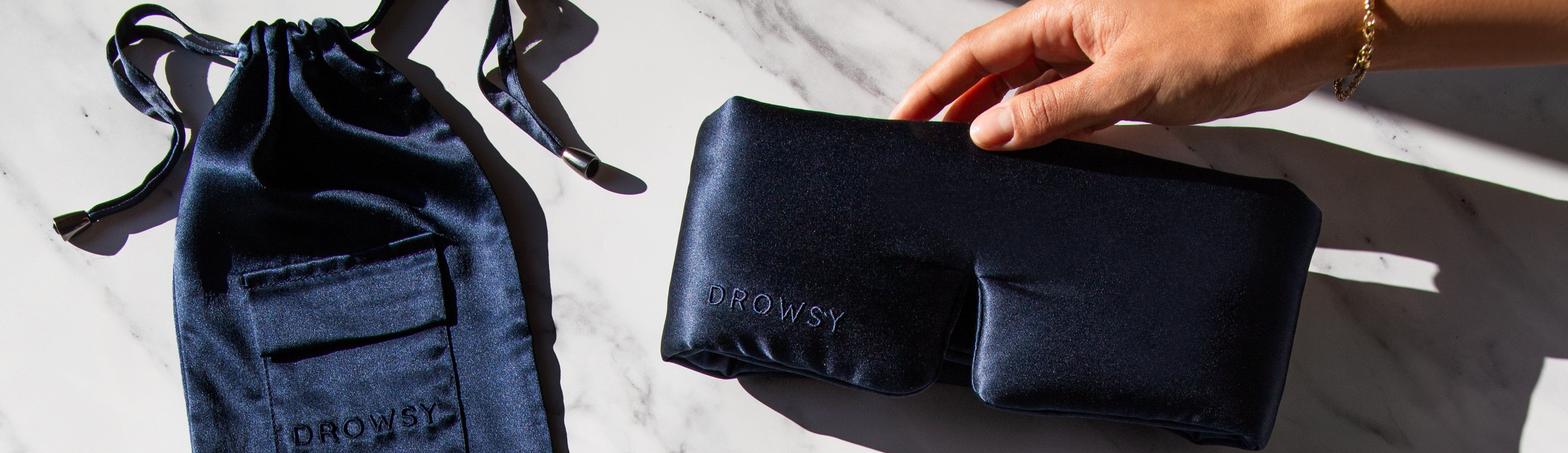 A hand holds a dark blue satin sleep mask with DROWSY embroidered on it, next to a matching satin drawstring pouch on a white marble surface.