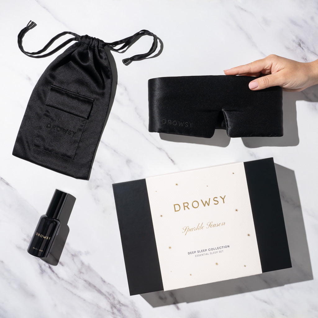A hand holds a luxury silk sleep mask beside a black satin pouch, deep sleep pillow spray, and a white drowsysleepco Black Jade Deep Sleep Collection gift box on a white marble surface.