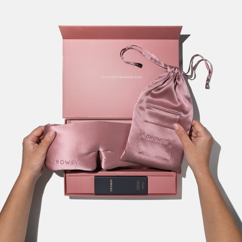 A person holds the drowsysleepco Damask Rose Deep Sleep Collection’s luxury silk sleep mask and matching pink pouch above an open pink gift box reading YOU DESERVE GLORIOUS SLEEP, with a dark deep sleep pillow spray box inside.