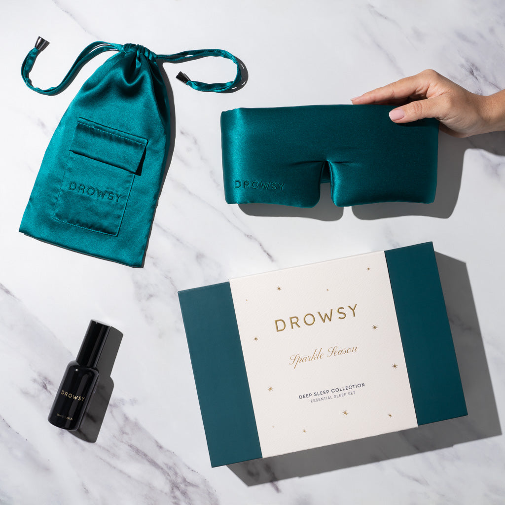 The Green Sapphire Deep Sleep Collection by drowsysleepco, featuring a silk sleep mask, green silk carry pouch, deep sleep pillow spray, and gift box, is displayed on white marble. A hand holds the eye mask.