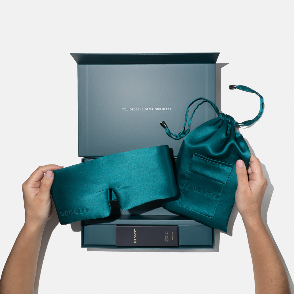 A person holds the drowsysleepco Green Sapphire Deep Sleep Collection silk mask and green pouch in front of an open gift box reading "YOU DESERVE GLORIOUS SLEEP," with a small product box inside, all set against a white background.