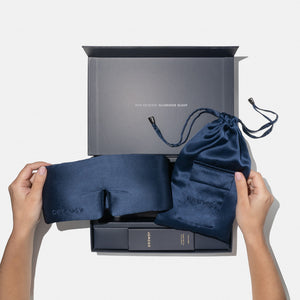 A person holds the drowsysleepco Midnight Blue Deep Sleep Collection silk sleep mask in front of an open gift box filled with matching essentials. "You deserve glorious sleep" is printed on the box.