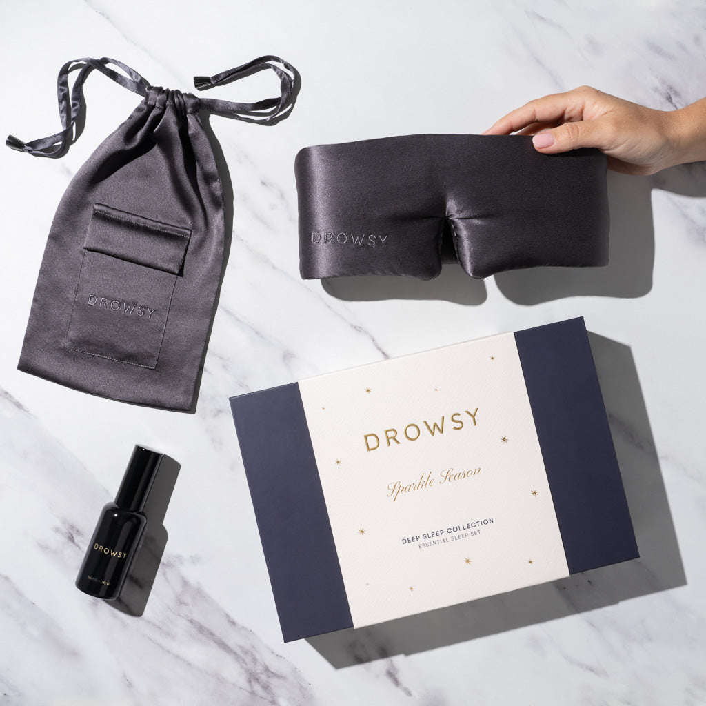 A hand holds a luxury silk sleep mask by drowsysleepco next to a matching pouch, black deep sleep pillow spray, and a boxed set labeled Moonlight Shadow Deep Sleep Collection on a marble surface.