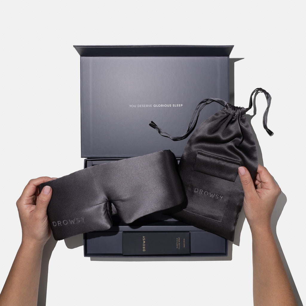 A person holds a drowsysleepco Moonlight Shadow Deep Sleep Collection silk mask and matching pouch above an open gift box reading YOU DESERVE GLORIOUS SLEEP, with a deep sleep pillow spray and other essentials inside.