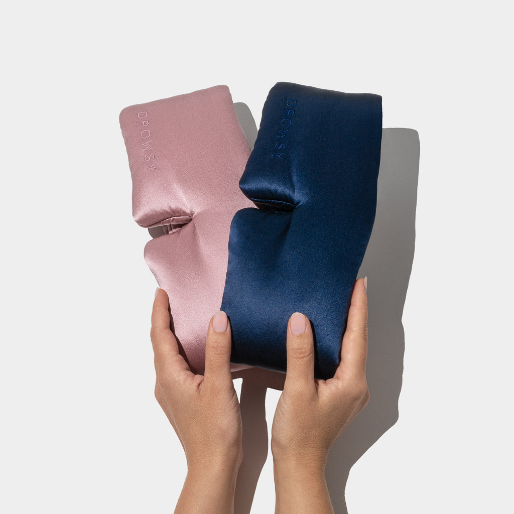 Two hands hold drowsysleepco’s Lovers Pack—two luxury silk sleep masks, one pink and one dark blue—against a white background, ideal for deep beauty sleep.