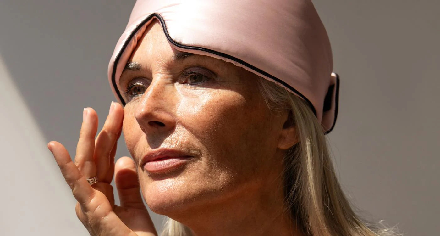 A woman with long gray hair wears a pale pink sleep mask pushed up on her forehead and gently touches her cheek, looking thoughtful in natural light.