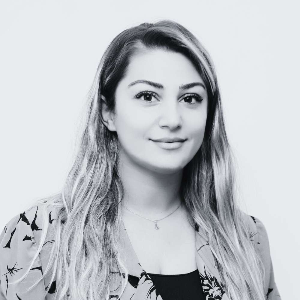 A woman with long, wavy hair wearing a patterned blazer and a necklace, looking at the camera and smiling slightly against a plain light background.