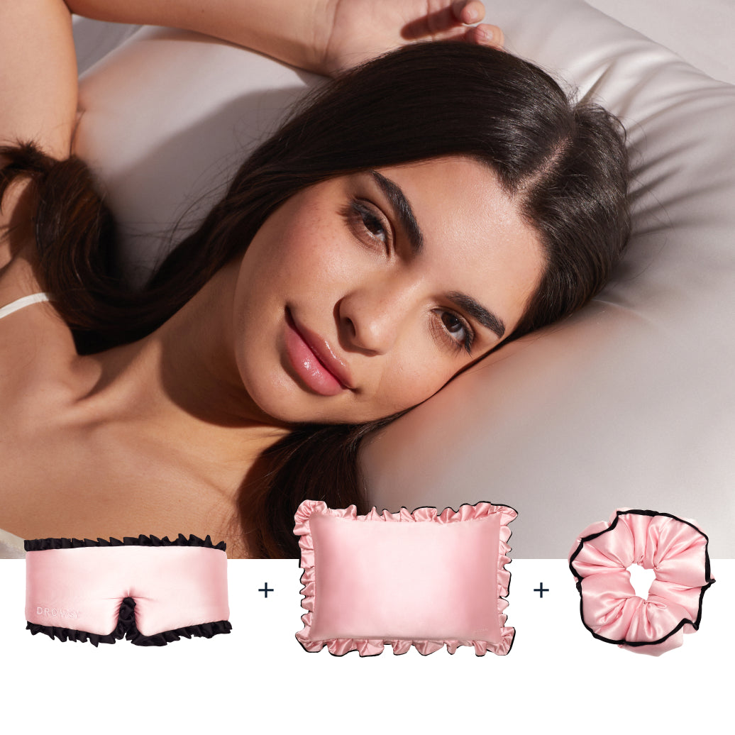 A woman with long dark hair lies on a satin pillow beside the drowsysleepco Manhattan Experience Bundle: a pink satin sleep mask with black trim, a ruffled pillow, and a scrunchie, all on a white background.