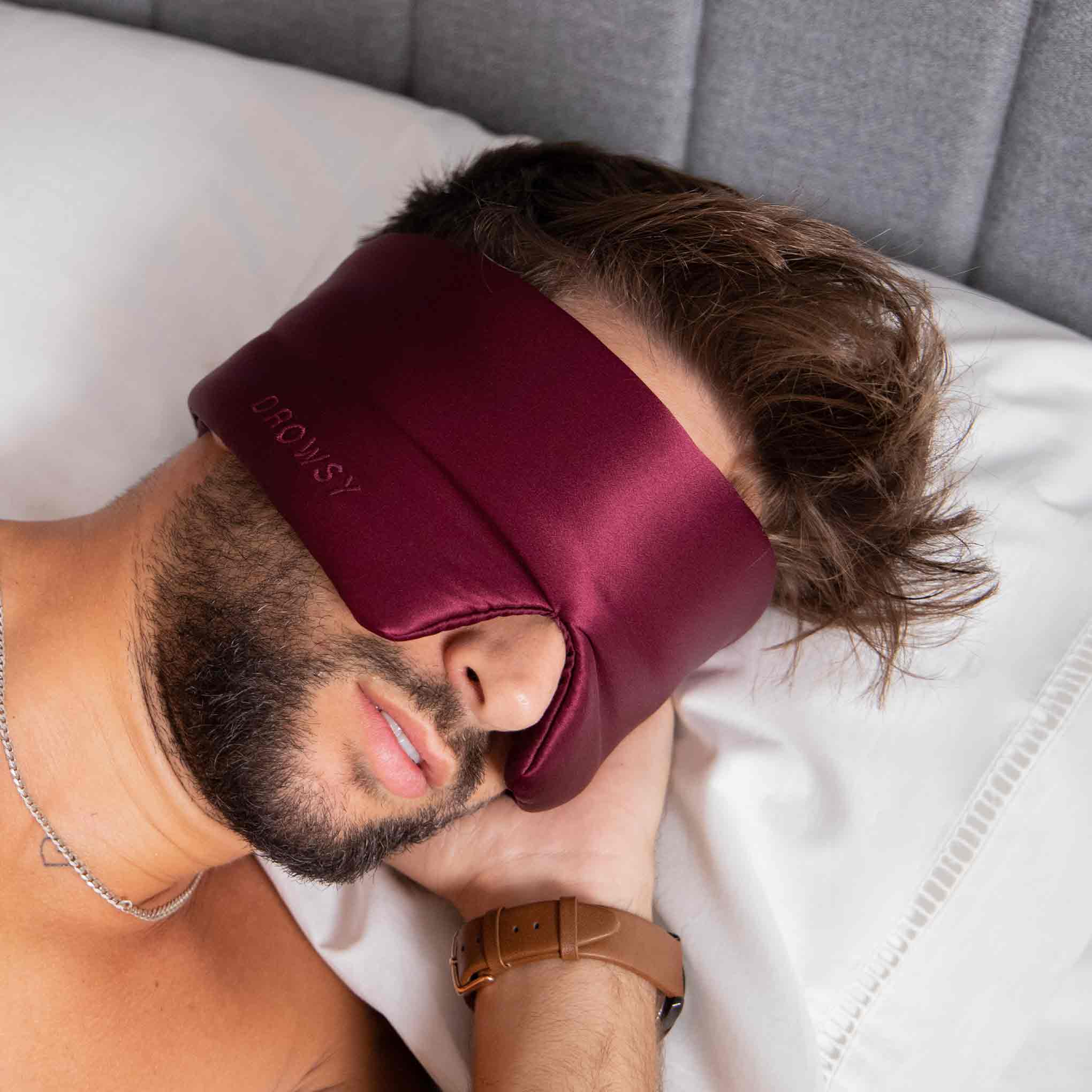 A man with a beard lies on his side in bed, wearing a burgundy satin sleep mask that covers his eyes and part of his face. He has tousled hair, a brown watch, and a thin silver necklace.