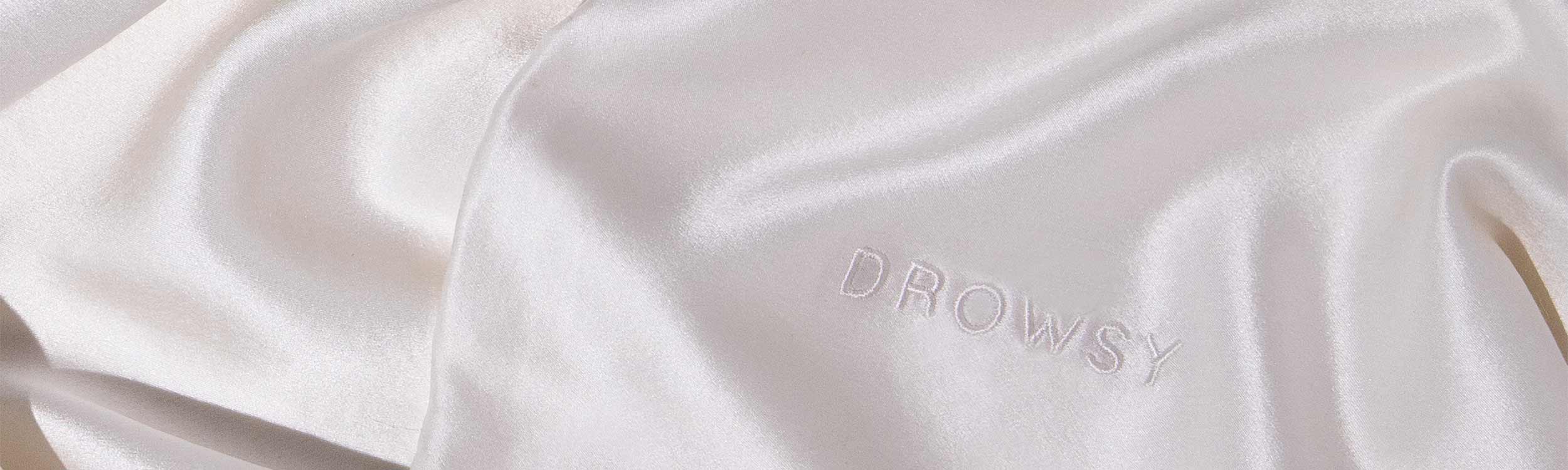 A close-up of smooth, light-colored silk fabric with the word DROWSY embroidered in matching thread. The surface appears shiny and soft, with gentle folds and highlights.