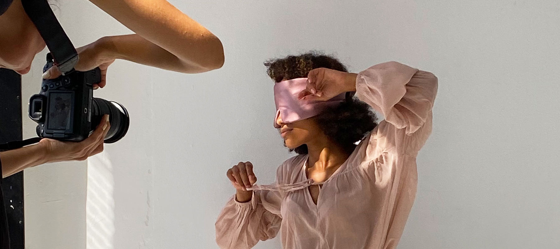 A photographer takes a close-up photo of a person wearing a light pink sleep mask and a matching loose blouse, posing against a plain white background.