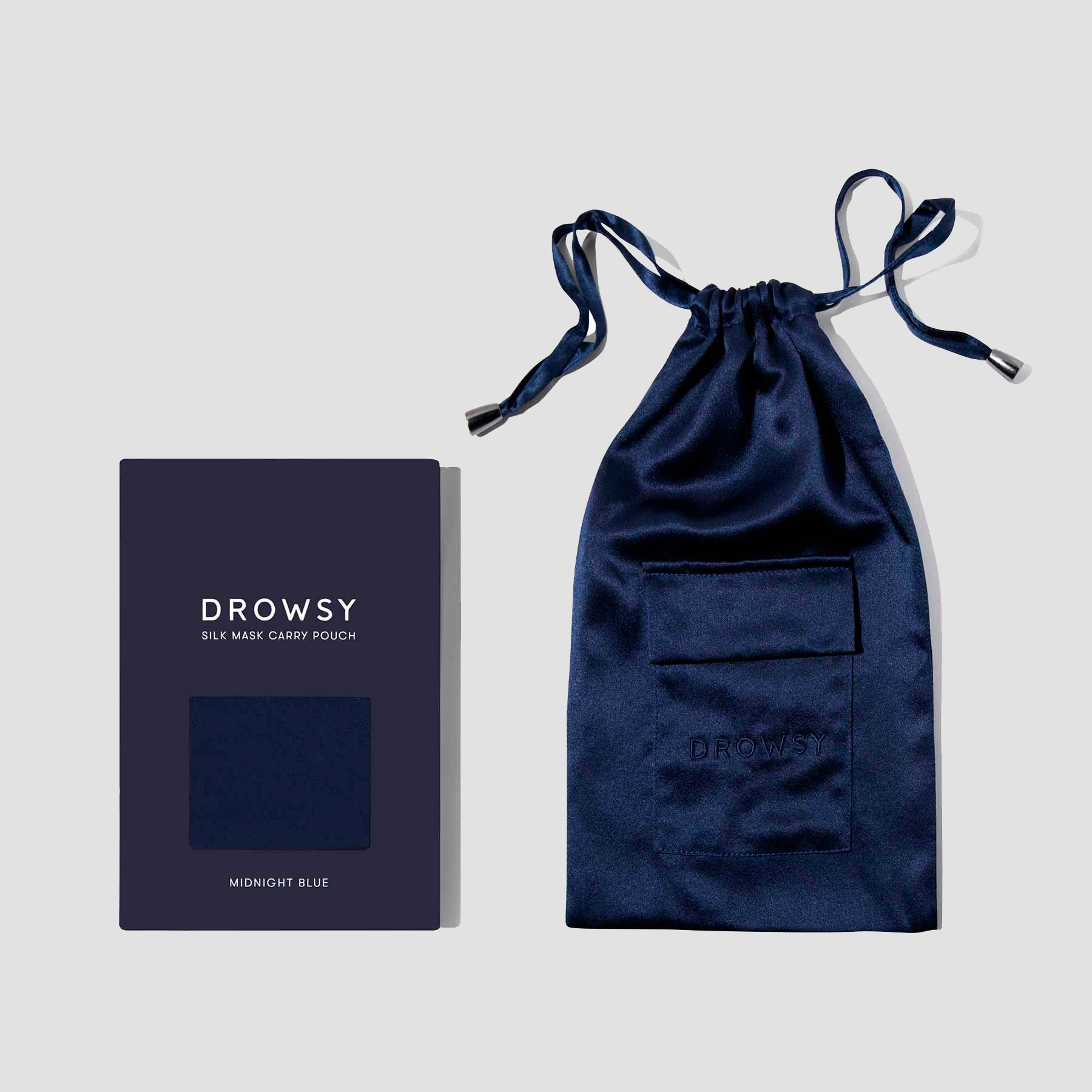A navy blue silk drawstring pouch with a small front pocket, placed next to its matching dark blue box labeled Drowsy Silk Mask Carry Pouch, Midnight Blue.