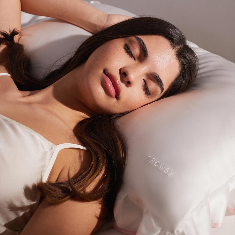 A woman with long brown hair sleeps peacefully on a white drowsysleepco Sleep and the City - Pearl & Pink Pillowcase, wearing a white satin camisole.