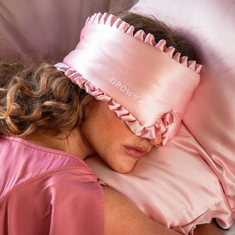 A person with wavy hair sleeps on the drowsysleepco Manhattan Experience Bundle, featuring a pink satin pillow, a pink ruffled satin sleep mask embroidered with DROWSY, and a matching pink satin top.