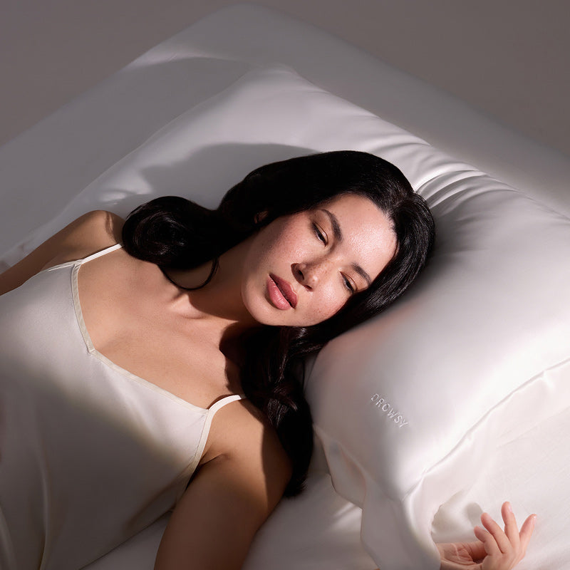 A woman with long dark hair sleeps peacefully on the us.drowsysleepco Hyalo Pillowcase - Akoya Pearl, dressed in a white satin camisole. Soft lighting accentuates her serene expression and the luxurious, smooth texture of the pillowcase.