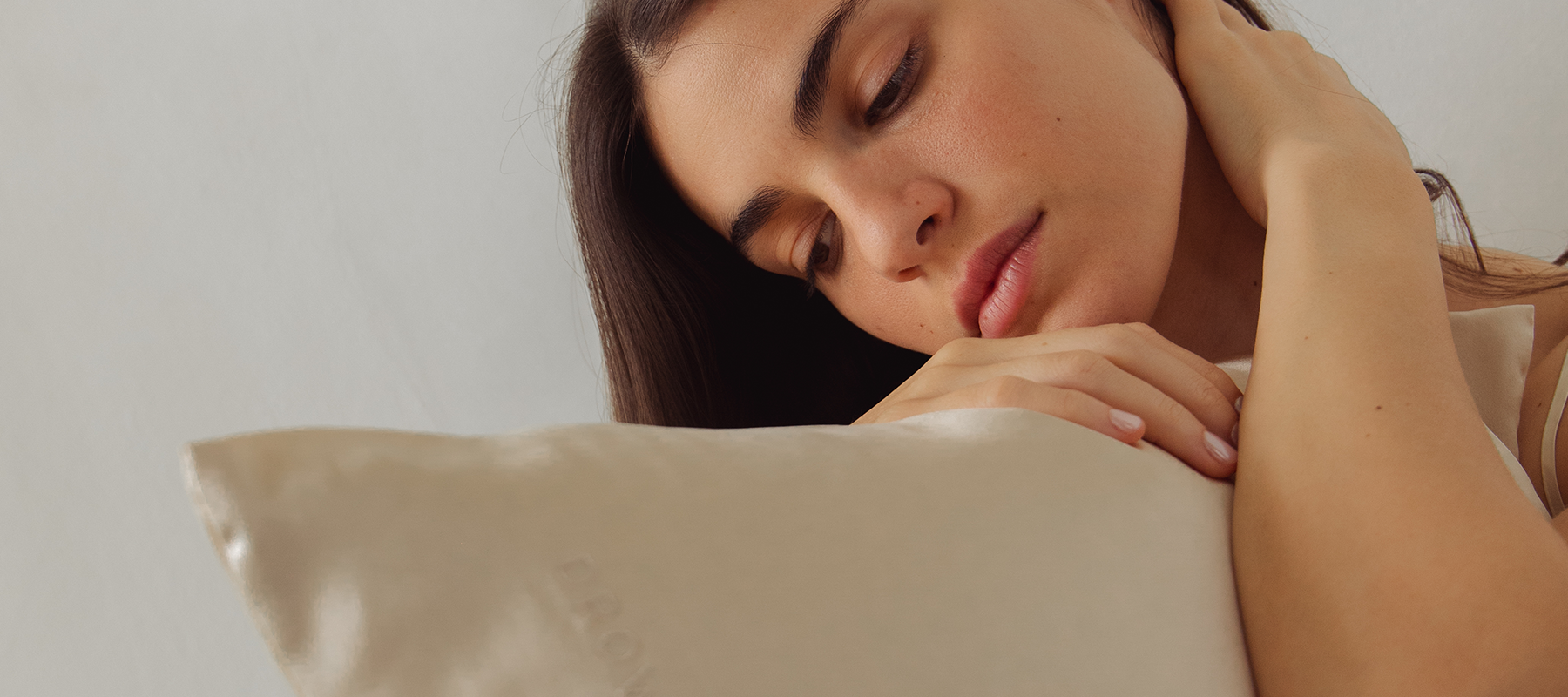 A young woman with long brown hair rests her head and hand on a light-colored satin pillow, looking down thoughtfully with a soft and relaxed expression against a neutral background.