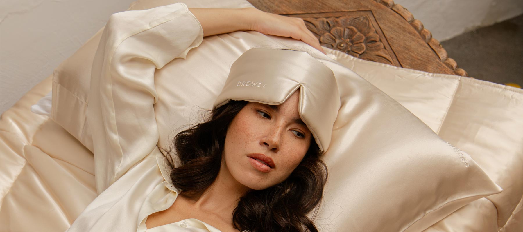 A woman with long dark hair lies on a bed with cream-colored satin sheets, wearing a matching satin sleep mask pushed up on her forehead and a satin shirt, looking relaxed with one arm resting on a carved wooden headboard.