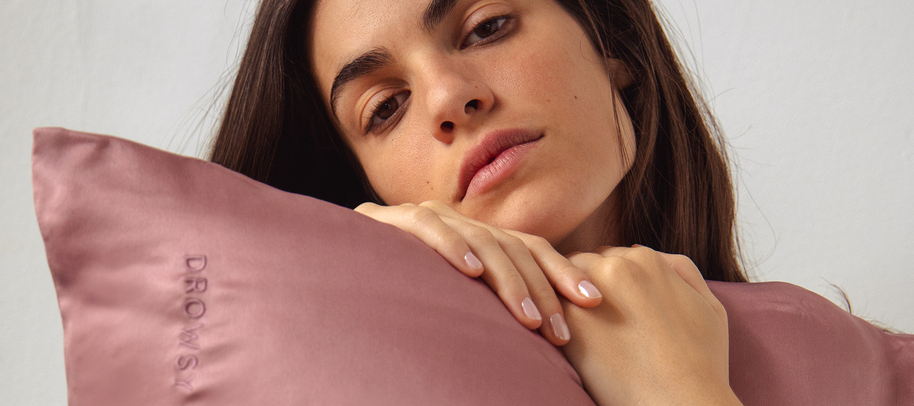 A woman with long brown hair rests her chin and hands on a mauve satin pillow embroidered with the word “DROWSY.” She has a calm, relaxed expression and wears natural makeup.