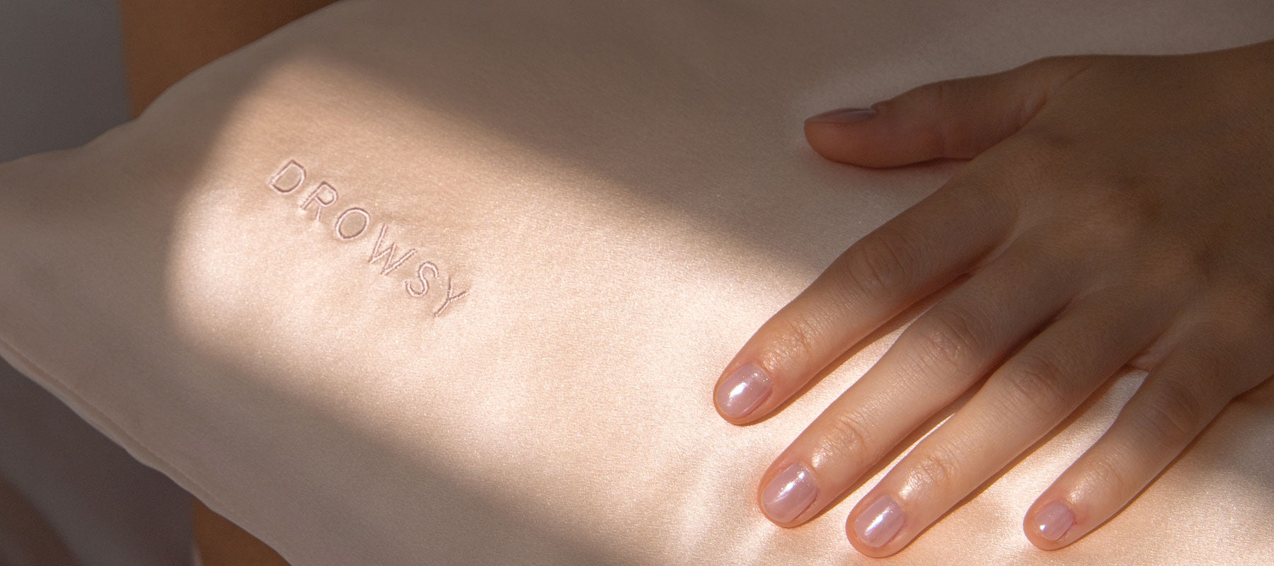 A hand with neatly manicured nails rests on a light pink silk pillow embroidered with the word DROWSY. Sunlight casts a soft shadow across the pillow.