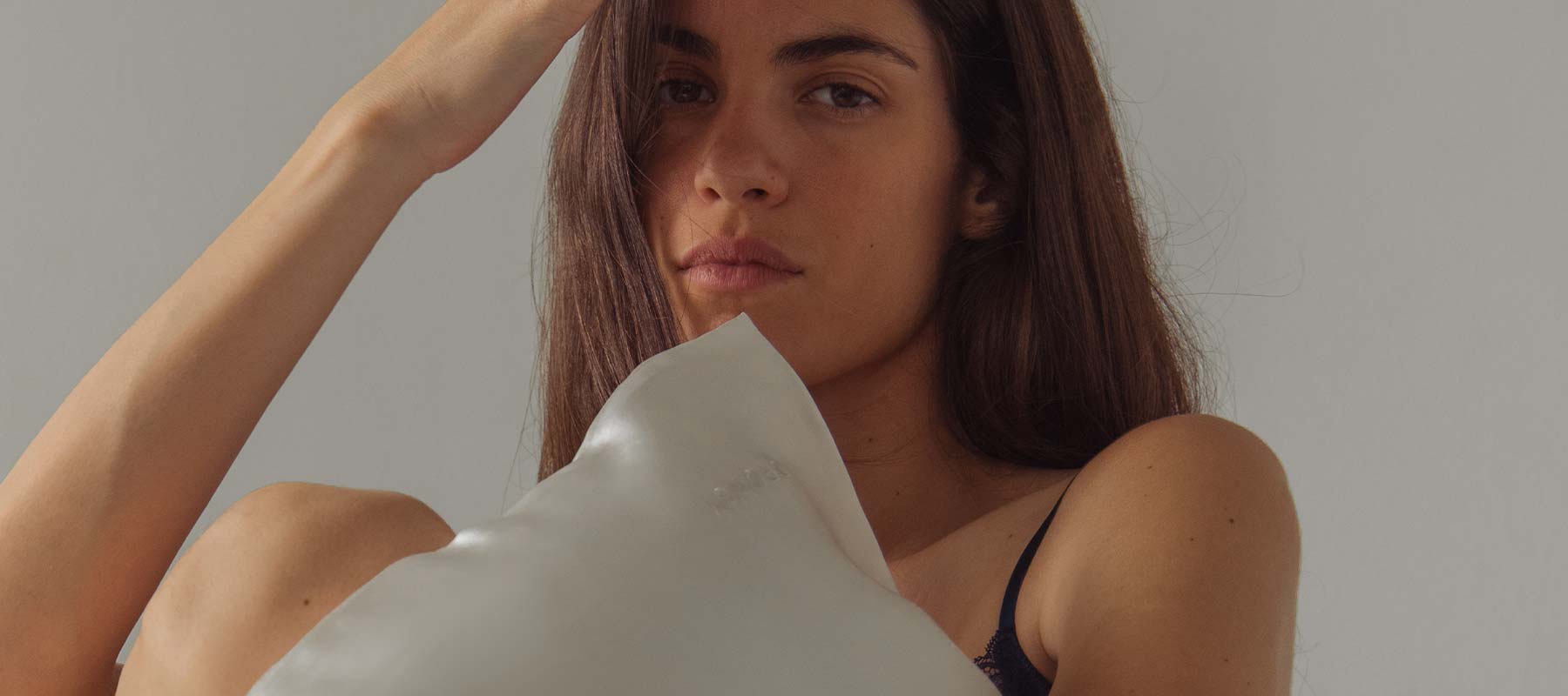 A woman with long brown hair holds a white pillow, looking directly at the camera with a neutral expression. She touches her head with one hand and wears a dark strap top. The background is plain and light-colored.