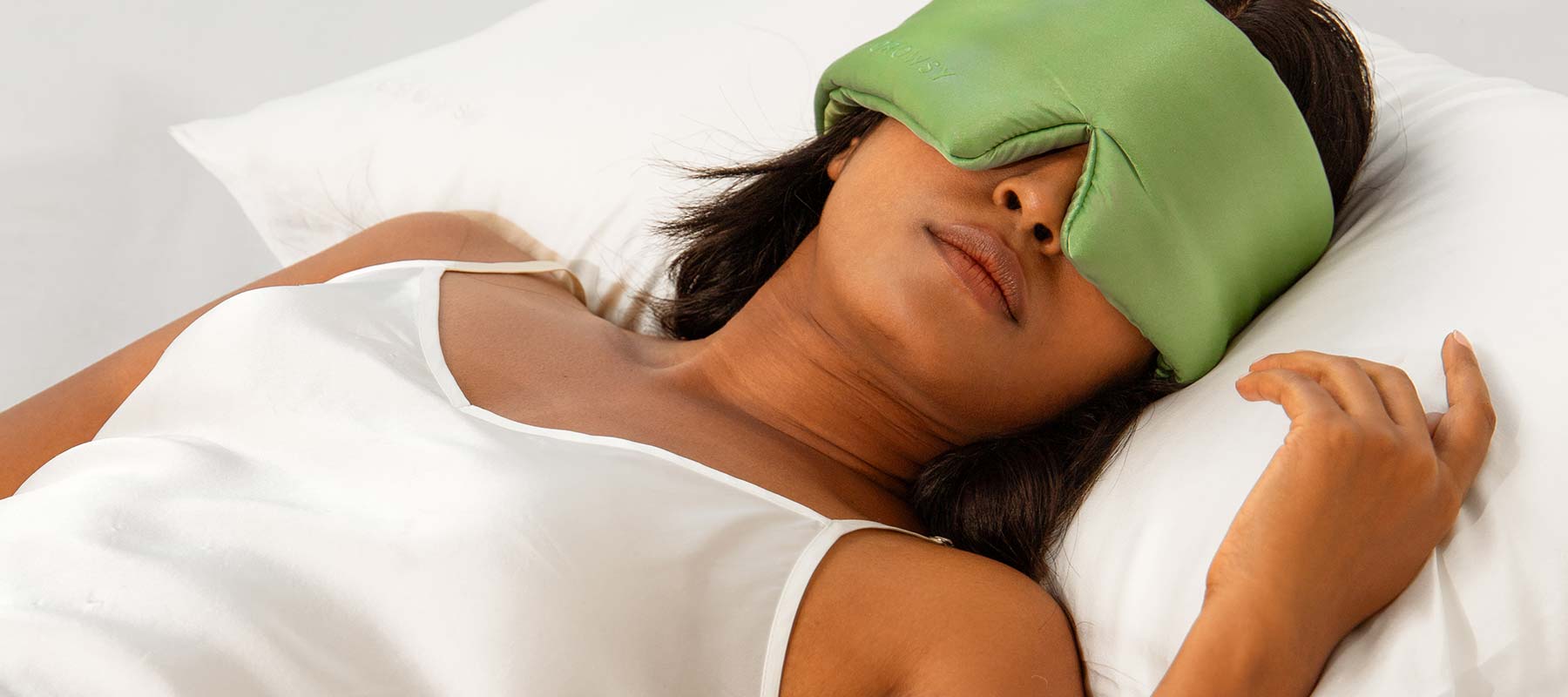 A woman in a white satin camisole lies on a white pillow and bed, wearing a large green eye mask, appearing relaxed and restful.