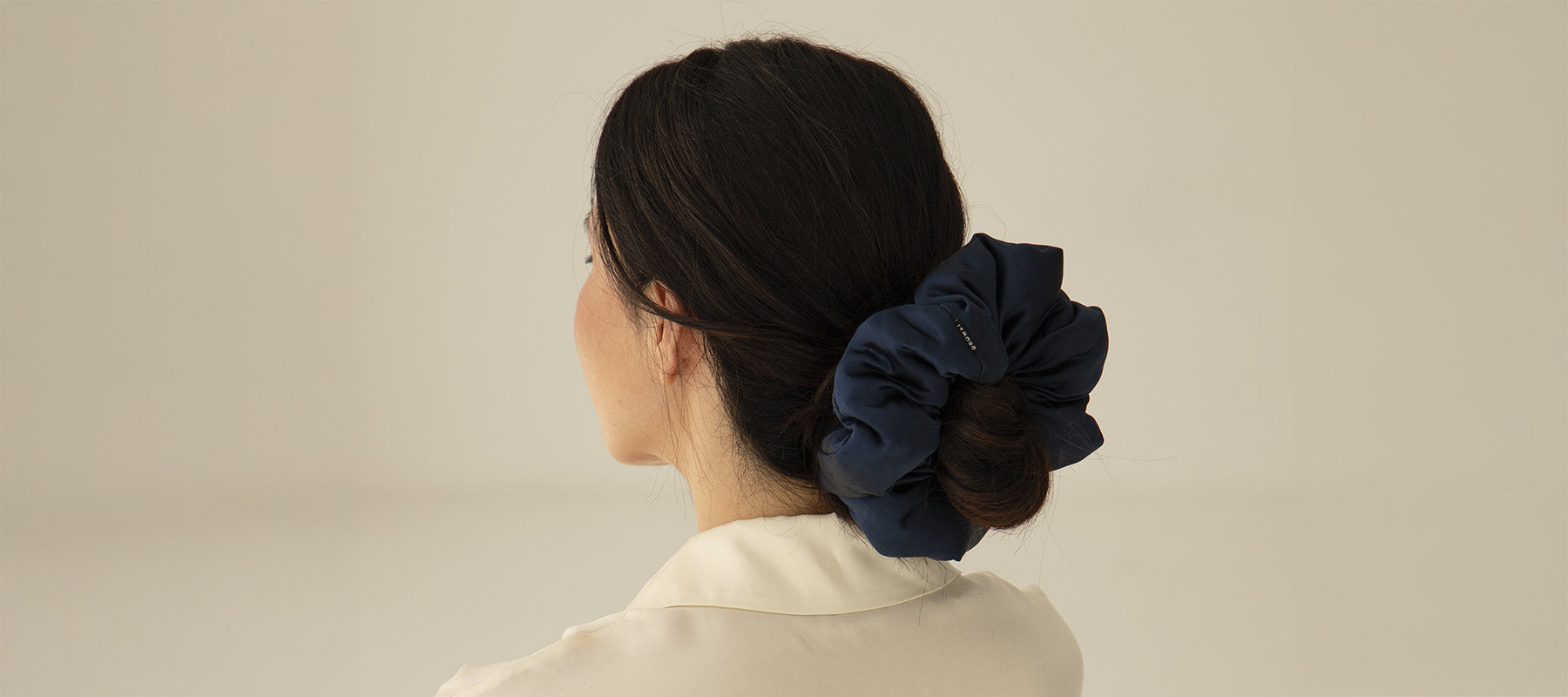 A woman with dark hair, seen from behind, wears a large navy blue scrunchie securing her low bun. She is dressed in a cream-colored top against a plain light background.