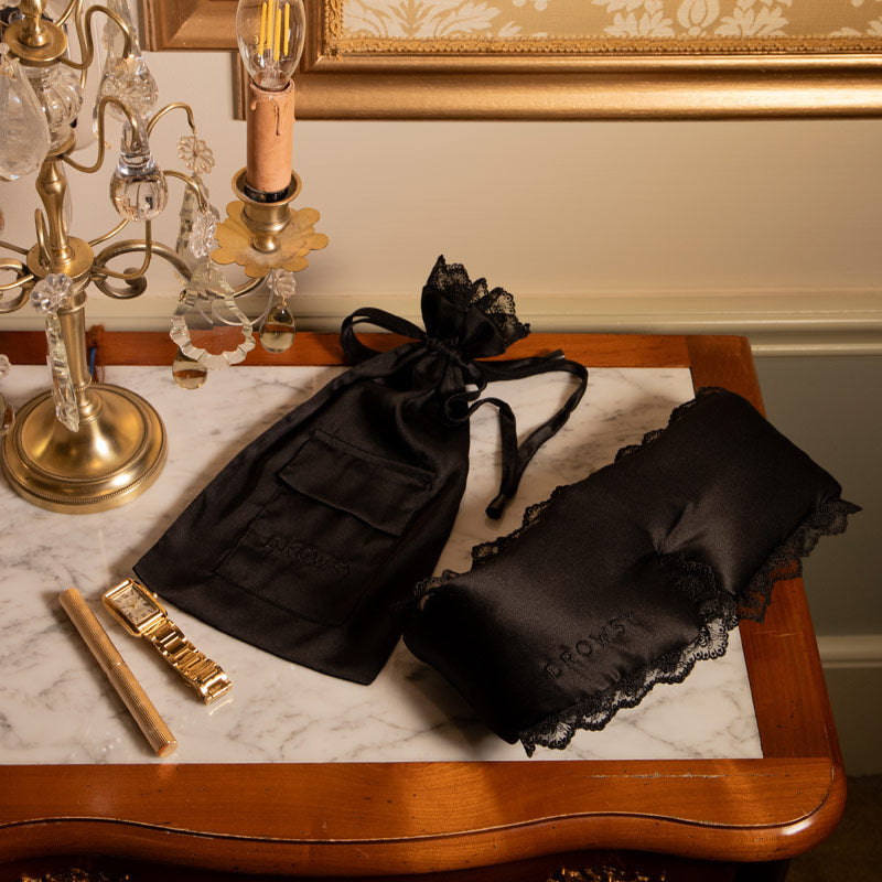 A marble-top table displays the drowsysleepco Paris Love Story - Black Jade Sleep Mask + Pouch, with lace trim and anti-aging benefits, beside a gold candle holder, gold comb, and an ornate lamp in warm light.