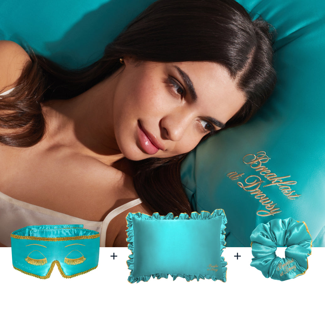 A woman with long dark hair rests on a turquoise satin pillow embroidered with "Breakfast at Drusy." In front is drowsysleepco’s Hollywood Experience Bundle: a turquoise silk sleep mask, ruffled pillow, and scrunchie for beauty sleep.