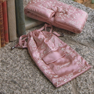 A pink mulberry silk Pride & Prejudice - English Rose Carry Pouch and matching sleep mask, both embroidered with white floral patterns and the word Drowsy, resting on a stone surface beside a row of antique books.