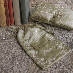 Pride & Prejudice - Sage Carry Pouch with a matching sleep mask, both featuring white floral embroidery and the word Drowsy, resting on a stone surface beside antique books.
