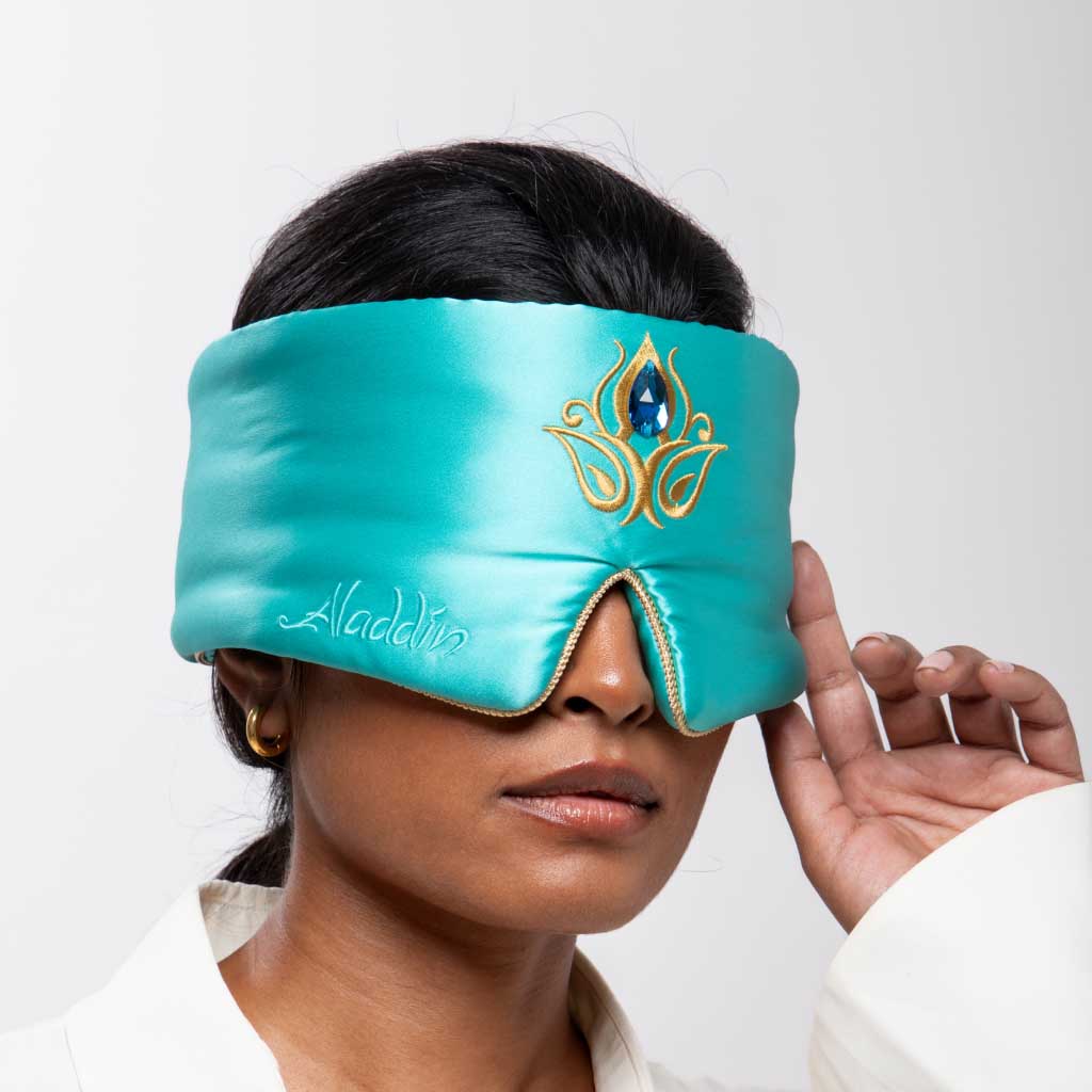 A person touches the edge of the Jasmine Jewel Sleep Mask by drowsysleepco, featuring teal mulberry silk with gold and blue embroidery and “Aladdin” stitched on the side, against a plain white background.