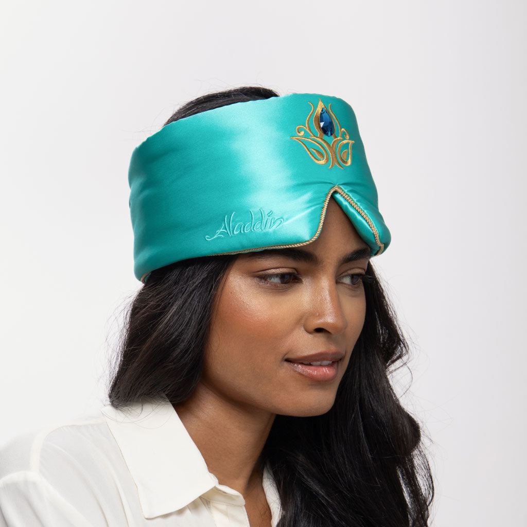 A woman with long dark hair wears the drowsysleepco Jasmine Jewel Sleep Mask, a turquoise mulberry silk mask embroidered with gold designs and 'Aladdin.' She is in a white shirt, looking to the side against a plain white background.