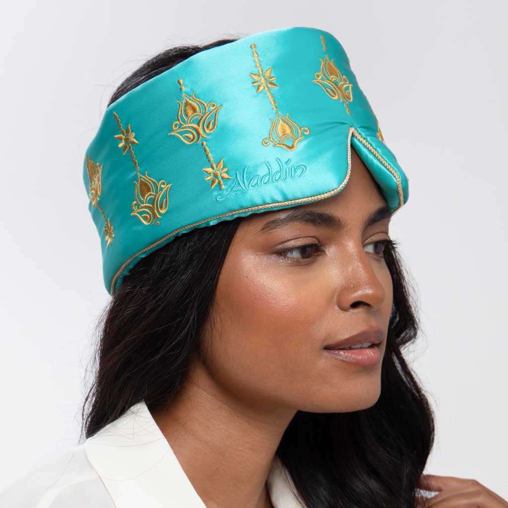 A woman with long dark hair wears the Jasmine Sleep Mask by drowsysleepco—a turquoise silk mask with gold designs and "Aladdin" embroidered on it. She faces slightly to the side and appears calm.