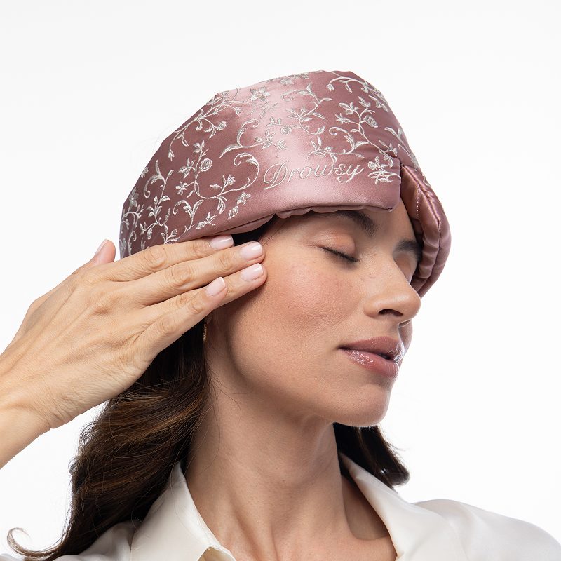 A person with closed eyes wears a mauve mulberry silk Pride & Prejudice - Sage + English Rose Eyelash Protecting Masks sleep mask, featuring white floral embroidery and the word Drowsy stitched in white thread.