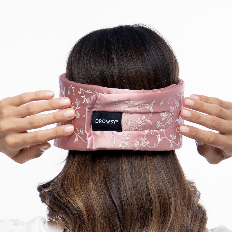 A person putting on a Pride & Prejudice - English Rose Eyelash Protecting Sleep Mask, a pink sleep mask with a white floral pattern and a black brand label on the back.