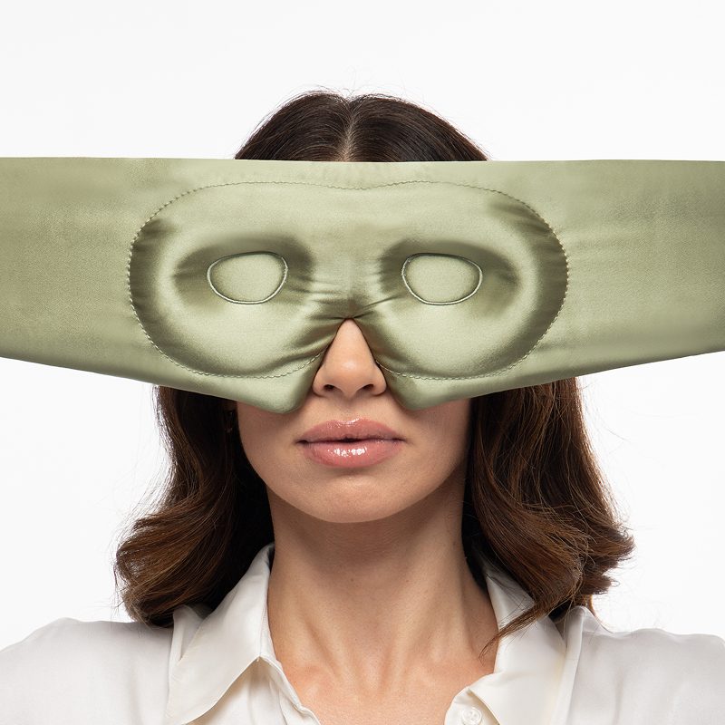 A person wearing a wide, sage-green Pride & Prejudice - Sage + English Rose Eyelash Protecting Masks silk sleep mask over their eyes.