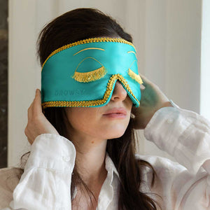 A woman in a white shirt puts on a drowsysleepco "2-Pack Sleep Masks | Breakfast at Drowsy," featuring turquoise fabric, gold trim, embroidered closed eyes, and gold fringe, while standing in a softly lit room.