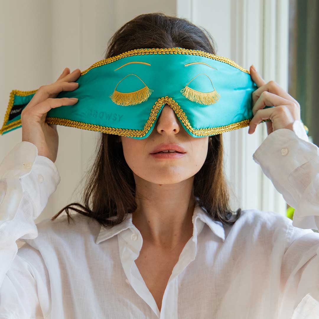 A woman in a white shirt puts on a turquoise sleep mask with gold trim and eyelash designs. She is standing indoors near a window, with natural light illuminating her face.
