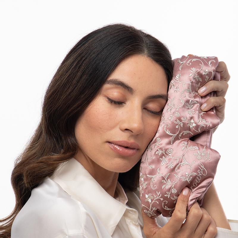 A woman with her eyes closed rests her cheek against a pink silk Pride & Prejudice - English Rose Sleep Mask embroidered with white floral vines.